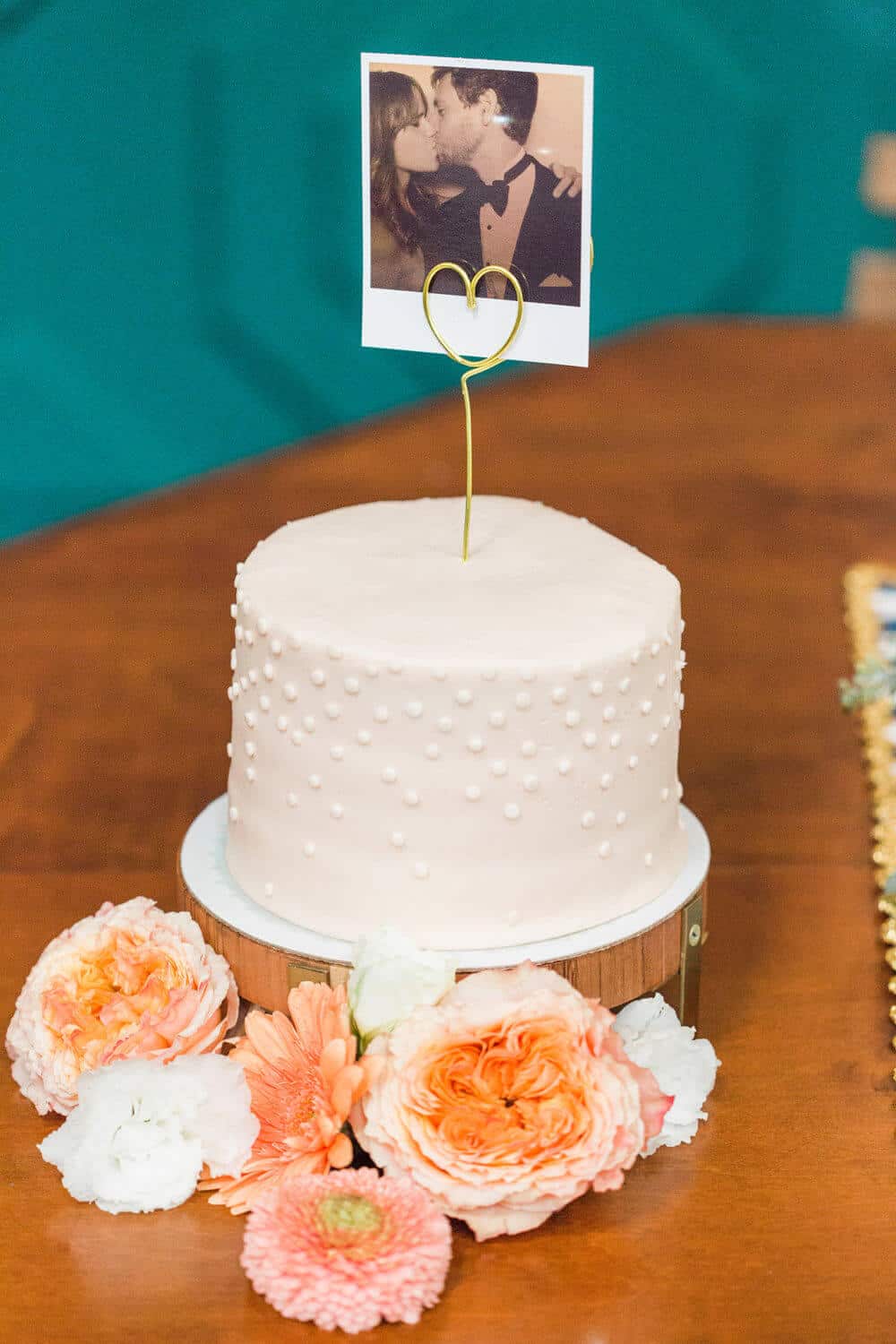 Petite wedding cake topped with a gold wire heart holder displaying a couple's photo, adorned with peach and white florals, set against a teal backdrop.