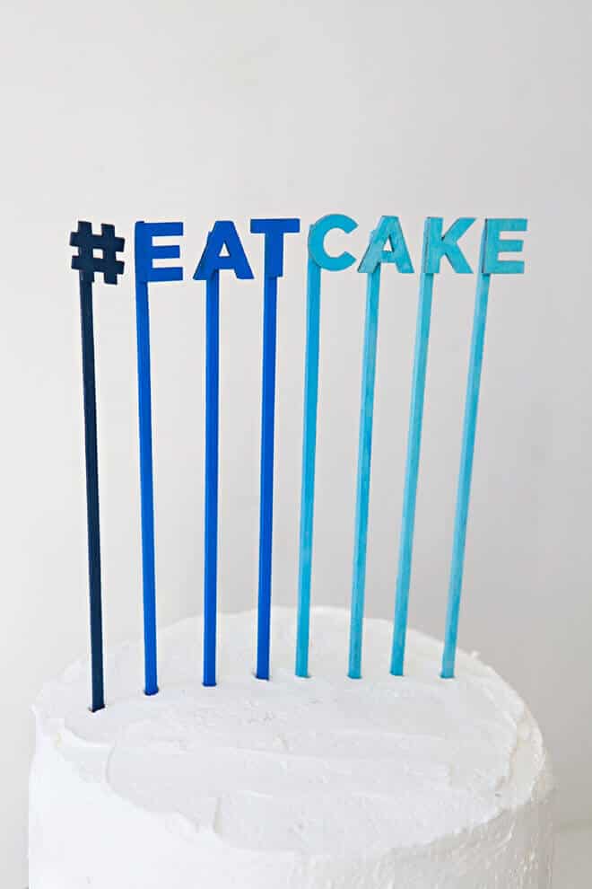 Minimalist white wedding cake topped with an ombré blue hashtag topper reading "#eatcake," perfect for modern celebrations.
