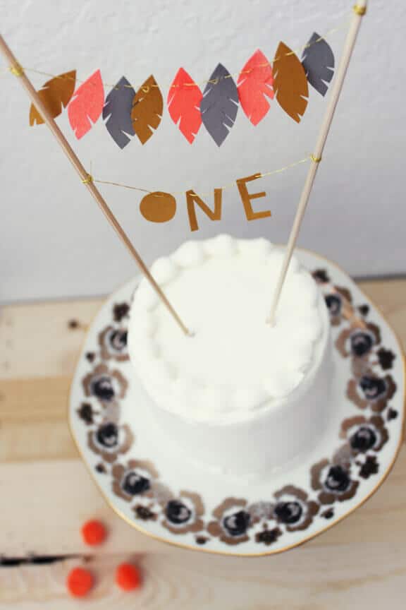 Single-tier white cake adorned with a paper feather garland and gold 'one' banner, resting on a floral-rimmed plate.