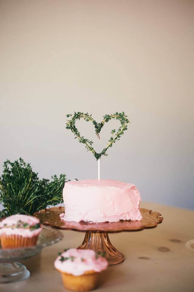 Blush buttercream wedding cake topped with a heart-shaped herb topper, displayed on an amber glass pedestal, with matching cupcakes nearby.