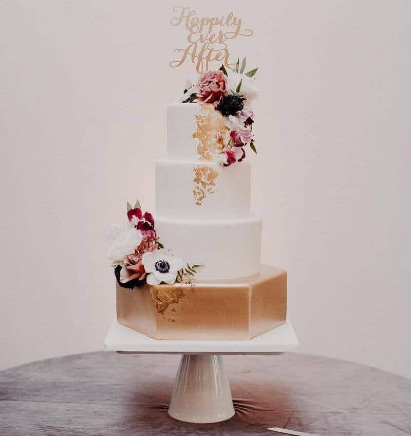 Three-tier wedding cake featuring a gold hexagonal base, white round tiers, gold leaf cascade, fresh burgundy and blush flowers, topped with "happily ever after. "