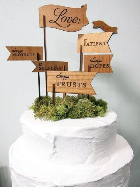 White buttercream wedding cake adorned with moss and rustic wooden arrow signs inscribed with virtues of love, perfect for a romantic setting.