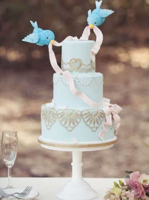 Powder-blue three-tier wedding cake with gold lace piping and two bluebirds holding a pink ribbon, set outdoors with floral accents.