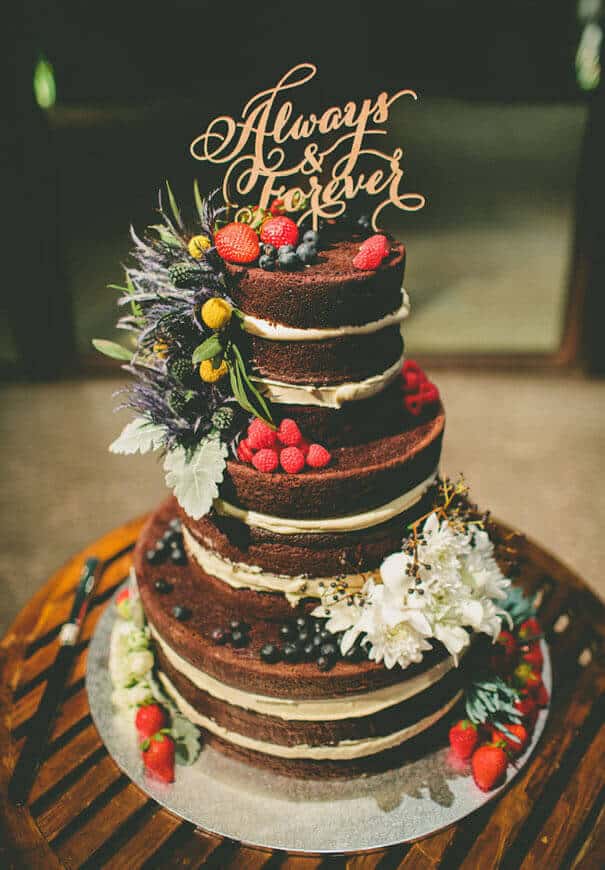 Three-tier naked chocolate wedding cake adorned with fresh berries and an 'always & forever' wooden topper on a rustic table.