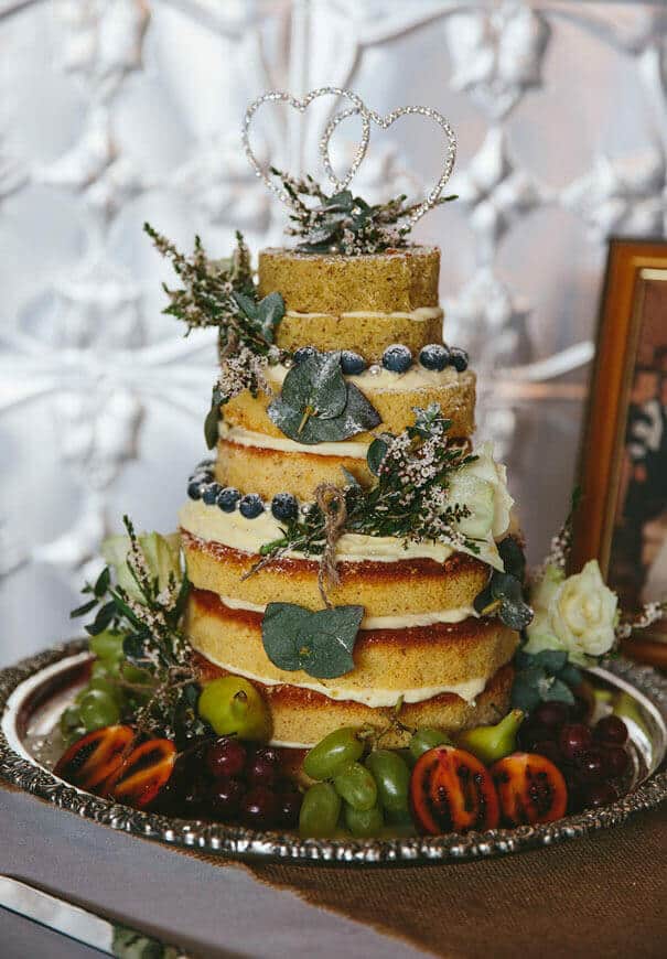 Rustic semi-naked wedding cake with a sparkling double-heart topper, adorned with blueberries, grapes, and eucalyptus, set against a pearl-silver backdrop.