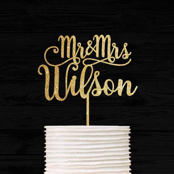 Elegant gold script 'mr & mrs wilson' cake topper on a white tiered cake, set against a black wood backdrop. Perfect for weddings.