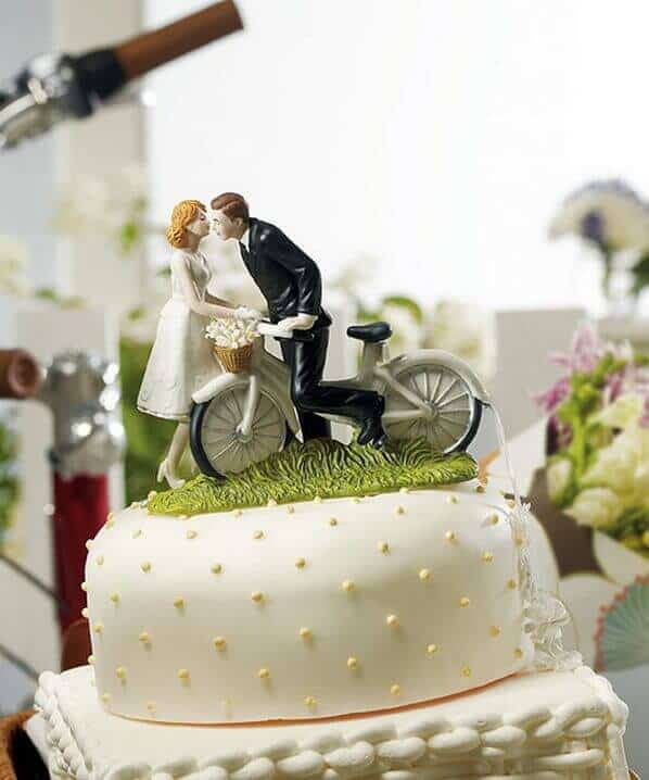 Ivory fondant wedding cake with a bride and groom figurine on a vintage bicycle, adorned with butter-yellow dot piping and floral backdrop.