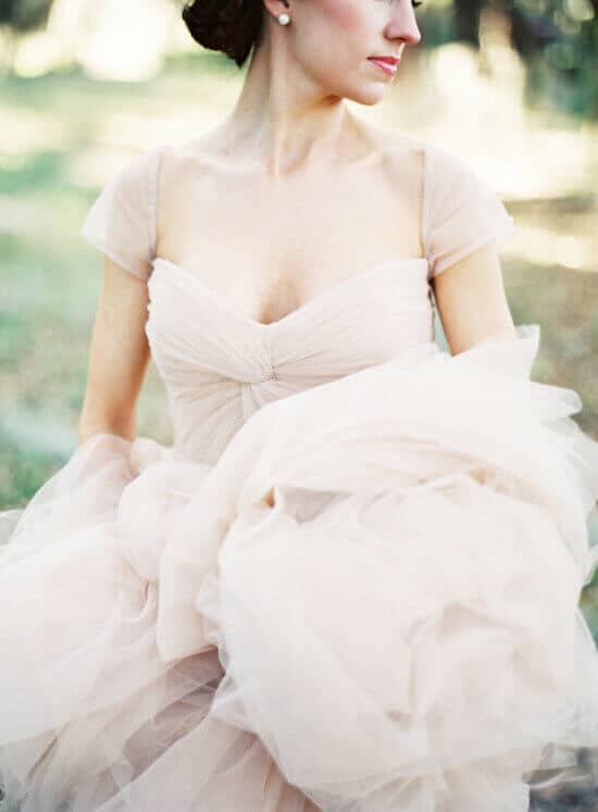 Blush tulle wedding gown with sheer cap sleeves, elegantly held by the bride in a garden, soft natural light highlighting its delicate layers.
