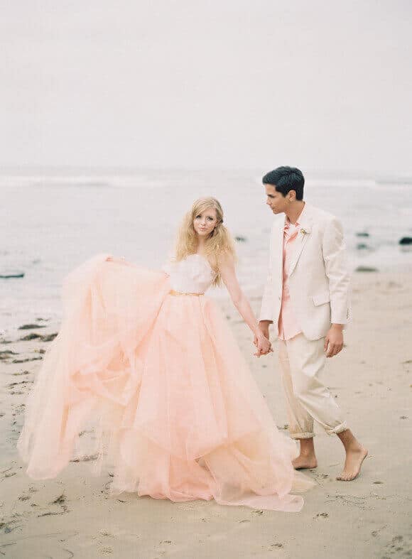 Bride in a flowing blush-peach tulle gown and groom in an ivory suit walk barefoot along a misty beach, creating a romantic scene.