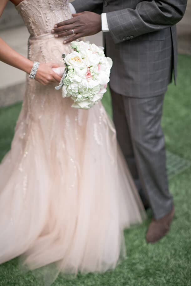 A bride in a blush-champagne tulle gown with beadwork, holding a bouquet of ivory and blush roses, stands on a manicured lawn beside her groom.