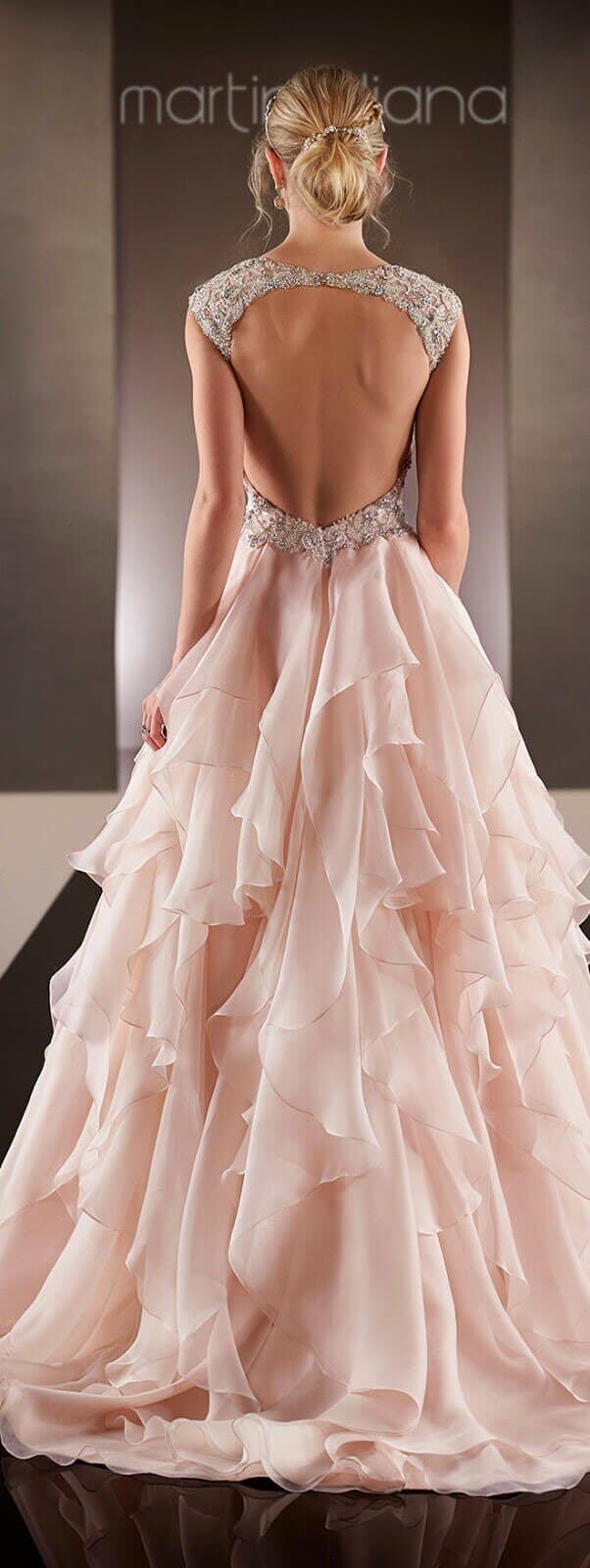 A back view of a blush pink wedding gown featuring a deep scoop open-back and beaded straps, with cascading organza ruffles creating a soft train.