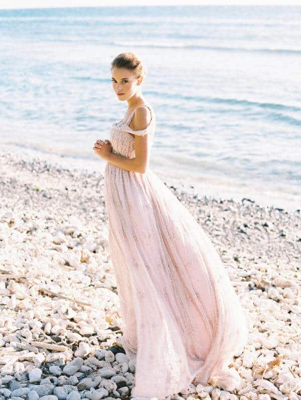 A woman in a flowing blush wedding gown stands on a pebble beach, sunlight illuminating the fabric as ocean waves gently lap the shore.