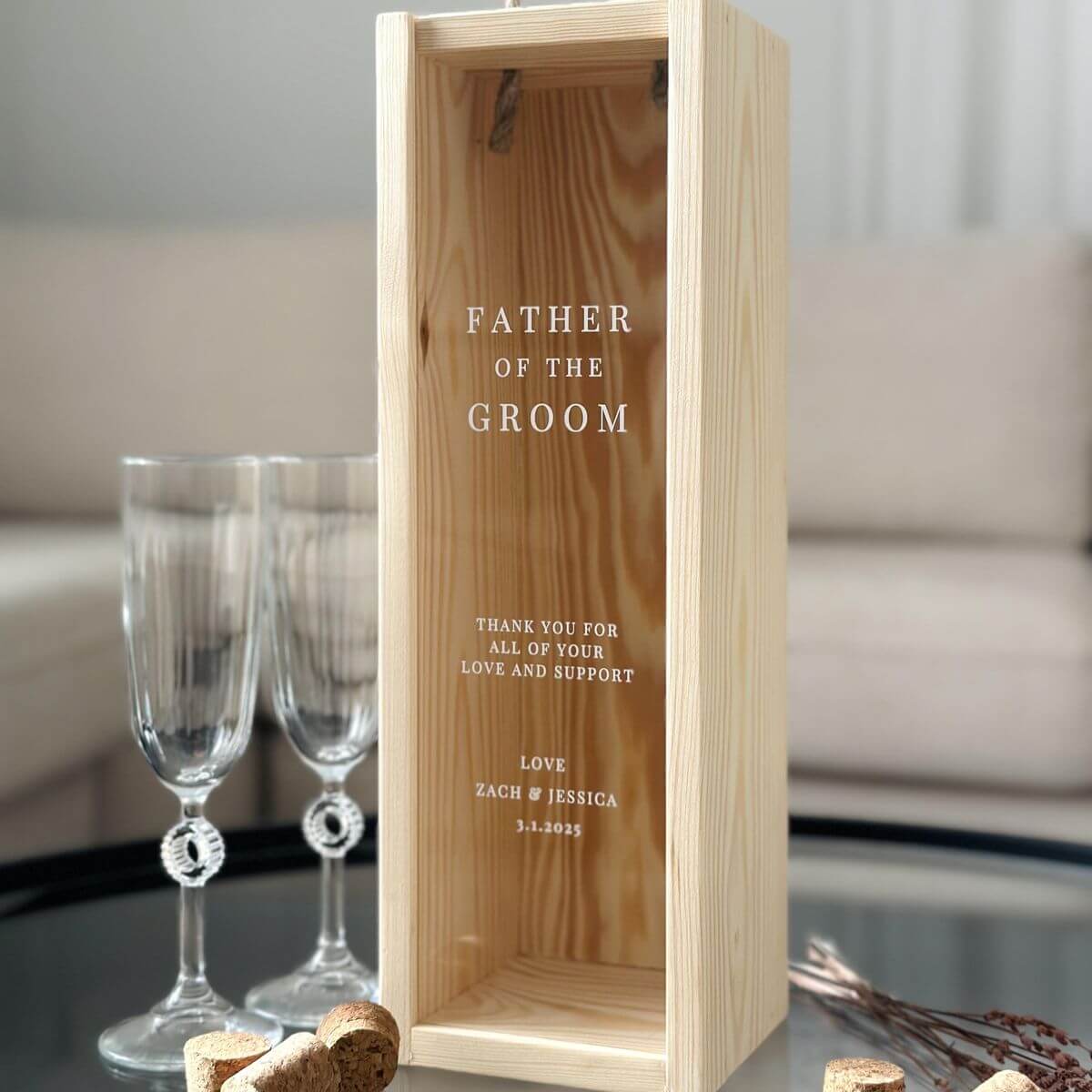 Personalized wooden wine box with engraved message for the father of the groom, accompanied by elegant champagne flutes on a glass table.
