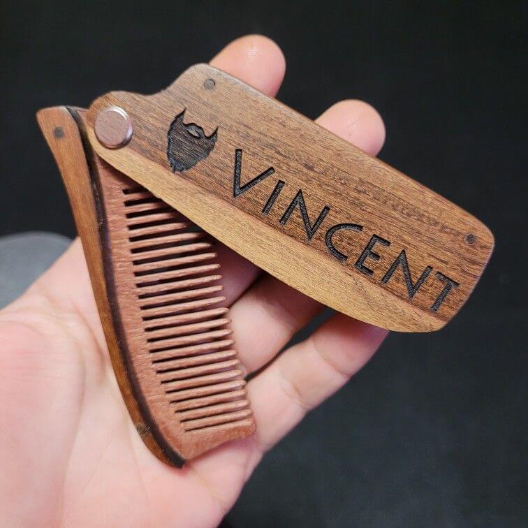 Personalized folding wooden beard comb with the name "VINCENT" engraved, ideal groomsmen gift. Handheld view against a dark background.
