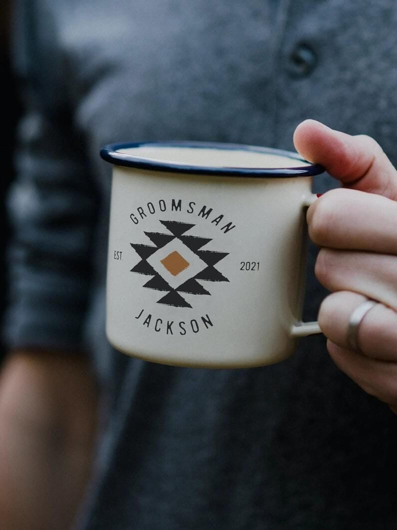 Close-up of a cream enamel camp mug with navy rim, reading "GROOMSMAN," personalized for Jackson, 2021, set against a blurred charcoal-gray henley.