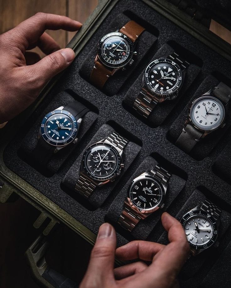 Close-up of eight wristwatches in an olive green hard case, hands selecting a timepiece, highlighting groomsmen gift ideas for a wedding.
