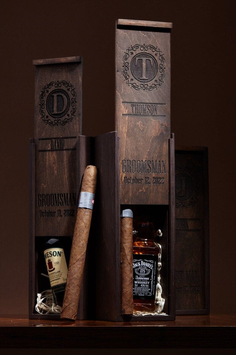 Three rustic dark walnut gift boxes, engraved with monograms, hold hand-rolled cigars and mini whiskey bottles, ideal groomsmen gifts.