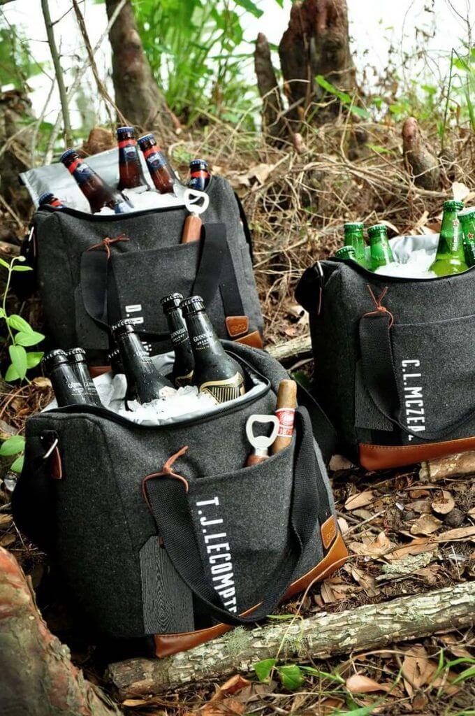 Three charcoal-gray monogrammed soft cooler bags with beer bottles in ice, nestled in a rustic woodland setting, ideal for groomsmen gifts.